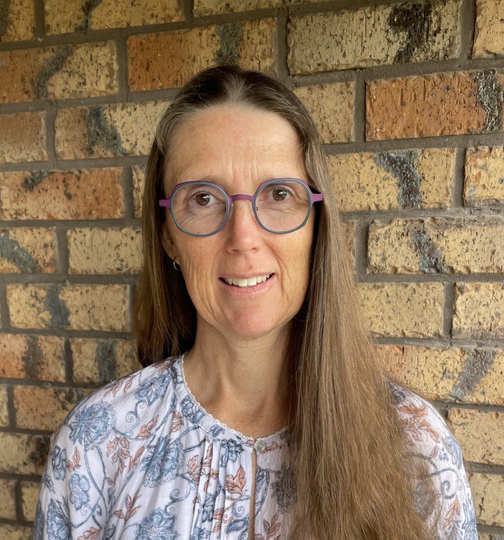 A woman with long hair wearing glasses standing in front of a brick wall.